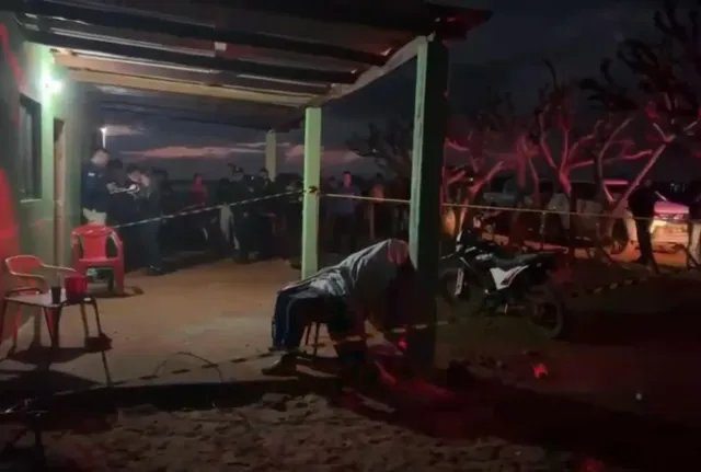 Cena do crime em frente a casa em Cerro Corá com fita policial, motocicleta e equipe de perícia recolhendo cápsulas