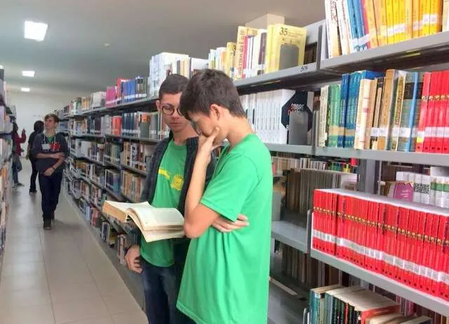 Pai consultando material escolar em biblioteca da rede estadual, com tablet exibindo audiobook e documento da lei nº 6.610 ao fundo