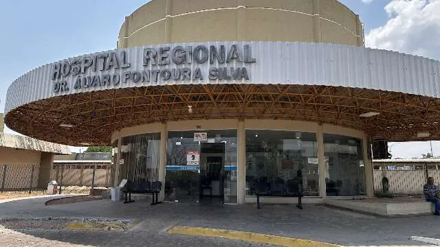 Bebê em observação em berço hospitalar enquanto equipe médica presta atendimento; mãe e policial ao fundo.