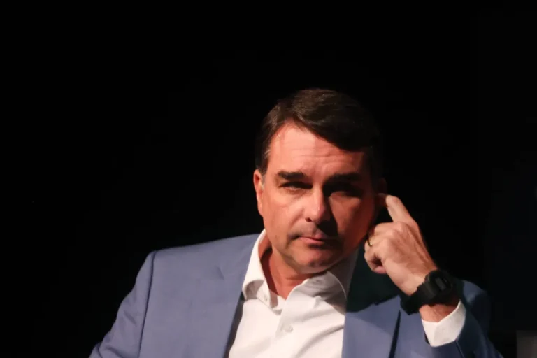 Flávio Bolsonaro em palco de convenção vazio com líderes do centrão em silhueta ao fundo