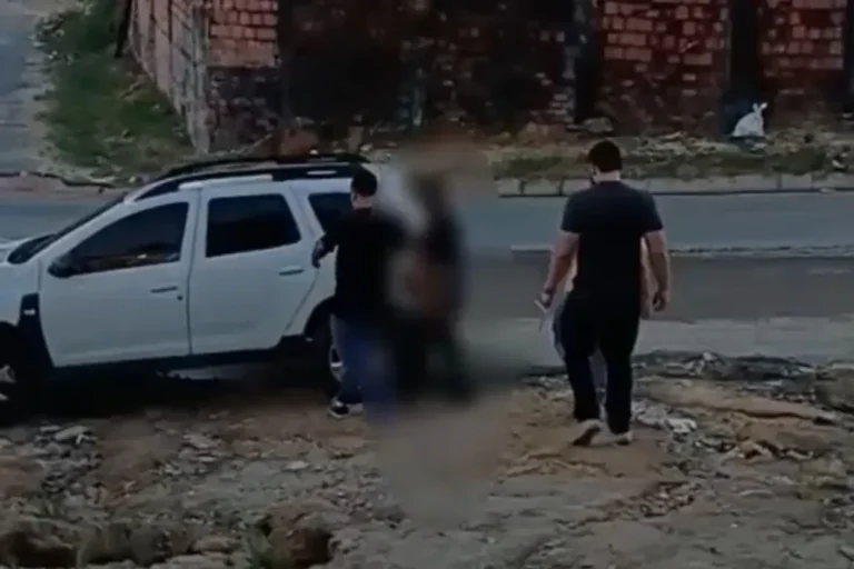 Fita de isolamento policial em frente a residência com viatura ao fundo, representando investigação policial.