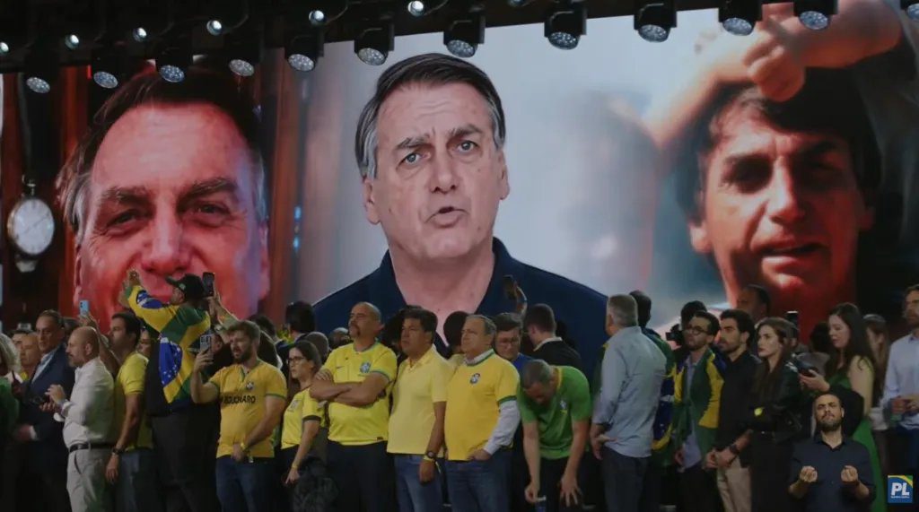 Flávio Bolsonaro discursando em convenção do PL com telão mostrando imagem digitalizada ao fundo