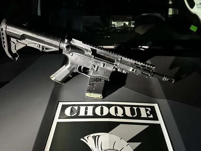Carabina M4 embrulhada no canteiro central de uma avenida com policiais e viatura ao fundo