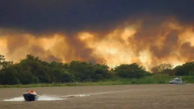 Área extensa queimada no Pantanal de Mato Grosso do Sul com coluna de fumaça e vegetação carbonizada; equipes de bombeiros atuando ao longe.