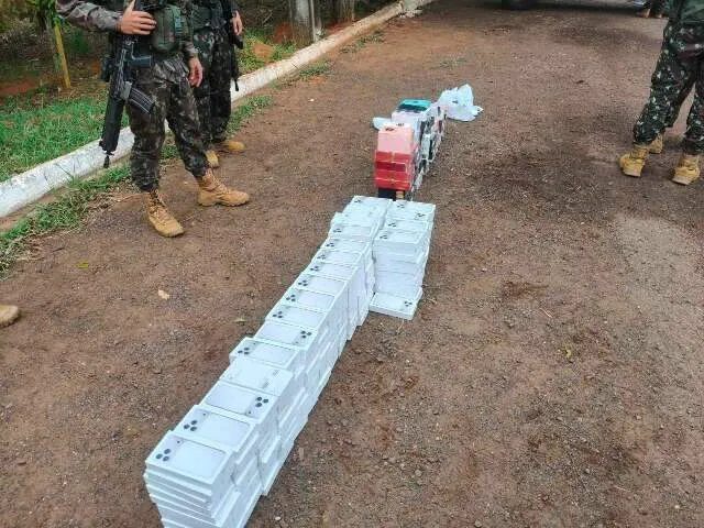 Carro com porta-malas aberto mostrando caixas de iPhones e frascos de perfume em posto fiscal, com policiais ao redor
